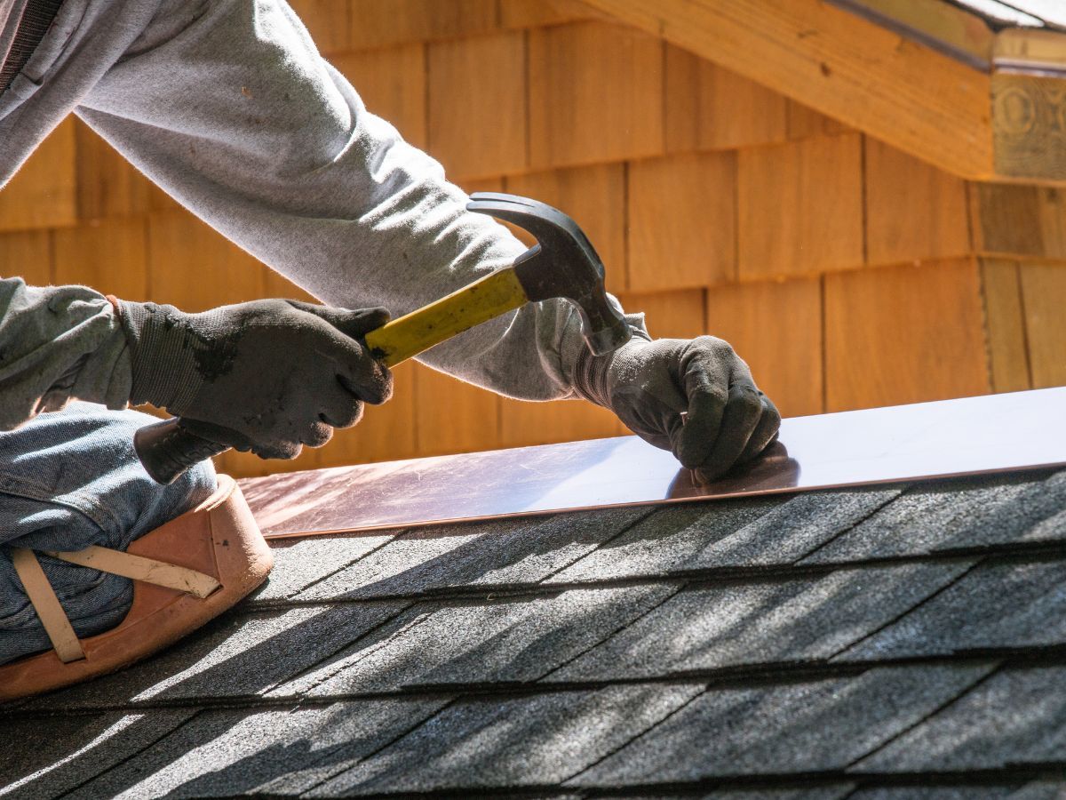 Roofer hammering metal flashing into place at the edge of a shingled roof